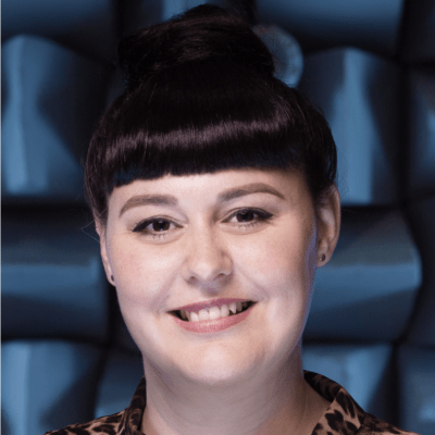 A profile picture of Kelly Miles. She has dark hair in a bun and a straight-cut fringe. She has dark eyes and is smiling. She is wearing a leopard-print blouse. The background is blue with a pattern resembling acoustic foam panels.