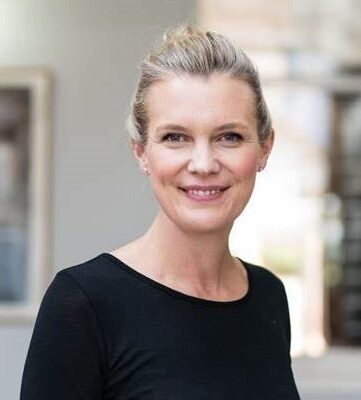 Professional headshot of a woman wearing a black top looking and smiling directly at the camera in front of a blurred background